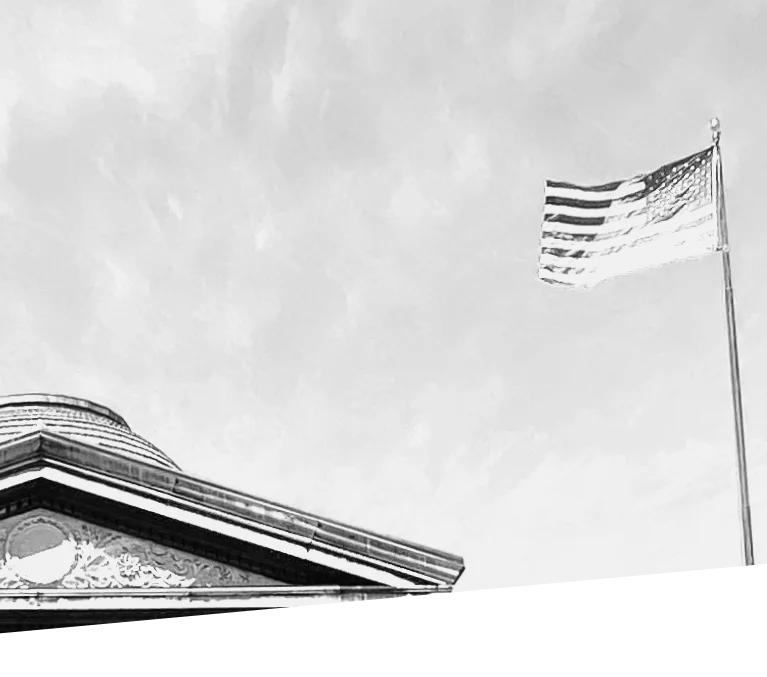 Black and white image of American flag waving on a flagpole next to a building with a triangular-shaped roof against a cloudy sky.