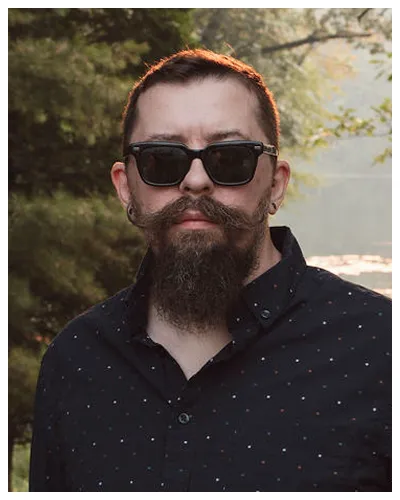 Man with dark beard, mustache, and sunglasses wearing a black shirt outdoors.