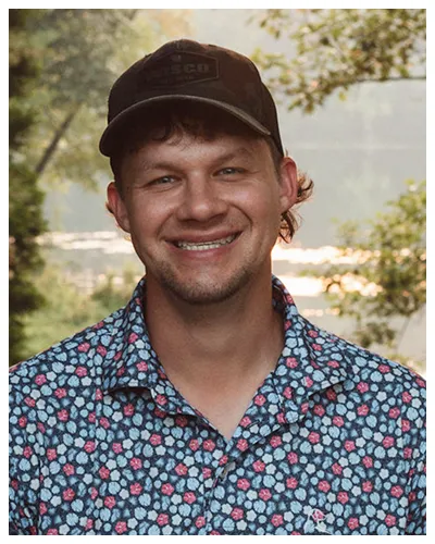Portrait of a smiling man wearing a black cap and floral patterned shirt outdoors.