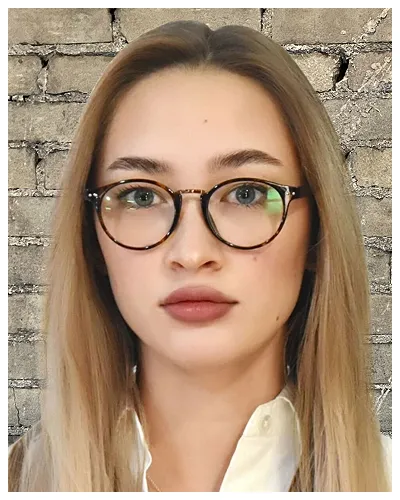Portrait of a woman with long blonde hair, wearing tortoiseshell glasses, white shirt, against a brick wall background.