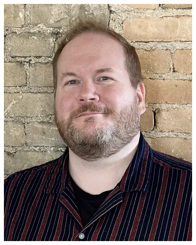 Portrait of a light-skinned man with short hair and beard, wearing a dark striped shirt, standing against a brick wall.