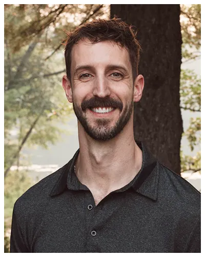 Portrait of a smiling man with short brown hair and beard, wearing a dark gray collared shirt, standing outdoors near a tree.