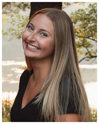 Portrait of a smiling young woman with long blonde hair, wearing a black top, outdoors near a lake.