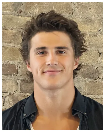 Young man with dark wavy hair wearing a dark shirt, standing against a brick wall.
