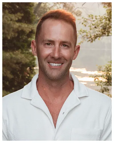 Portrait of a smiling man with short light brown hair wearing a white shirt outdoors.