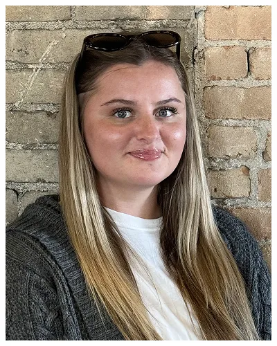 Portrait of a young woman with long blonde hair, wearing sunglasses on her head, white shirt, and gray sweater, against a brick wall background.