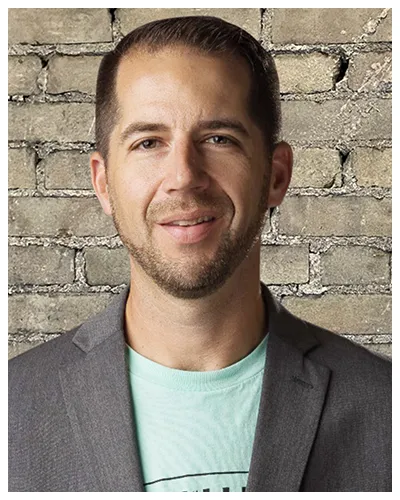 Portrait of a smiling man wearing a gray blazer and light green shirt against a brick wall.