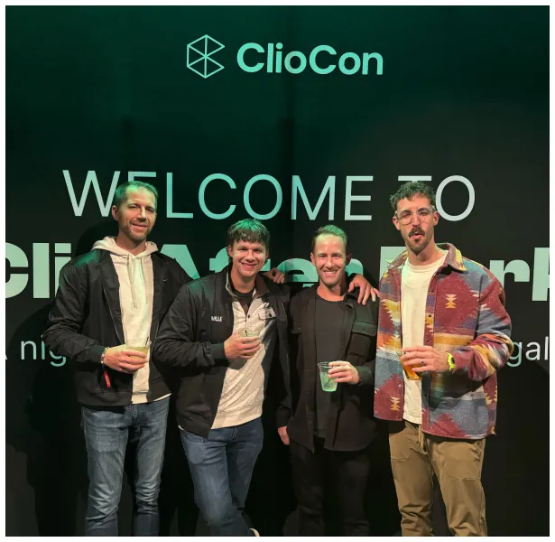 Four casually dressed men standing and holding drinks in front of a black backdrop with white text reading "ClioCon WELCOME TO".