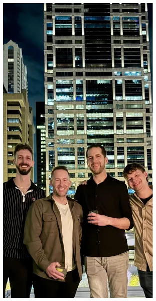 Four men smiling and posing outdoors at night with illuminated high-rise buildings in the background.