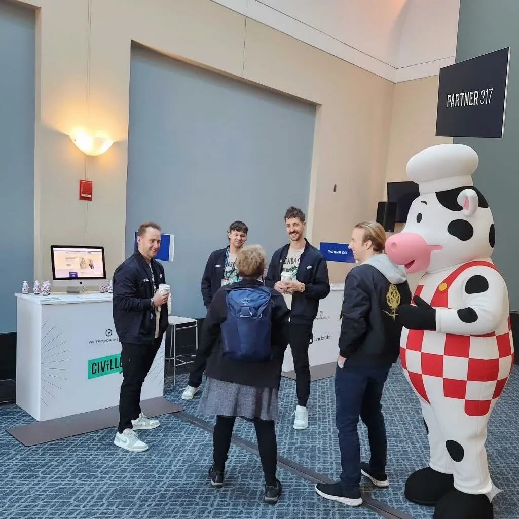 Group of five people conversing near a booth with computer and a large cow mascot in chef attire.