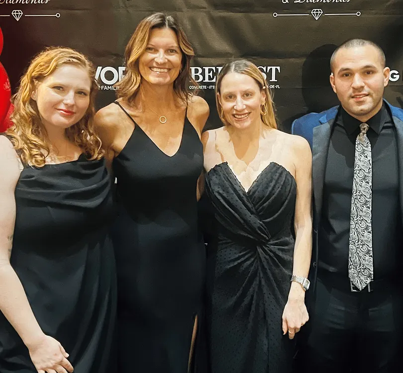 Four individuals in formal black attire posing in front of a branded black backdrop.