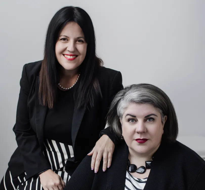 Two professionally dressed women, one standing with hand on seated woman's shoulder, both wearing black and white attire against a gray background.