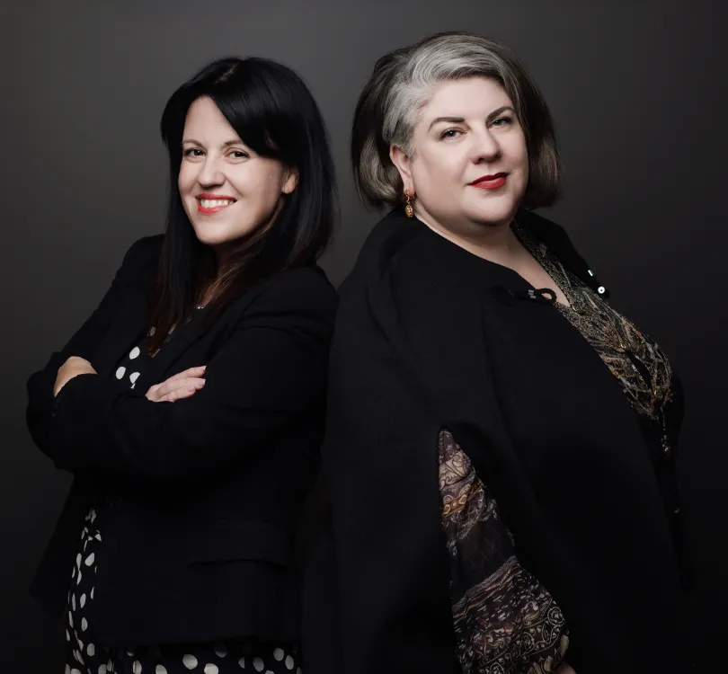 Portrait of two professional women standing back to back against a dark background, both wearing black attire.