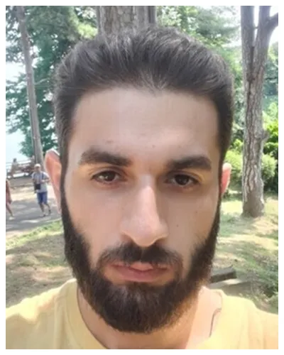 Close-up of a bearded man with dark hair outdoors in a wooded park area.
