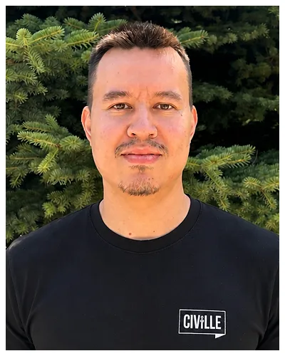 Portrait of a man with short hair wearing a black Civille shirt, standing outdoors near green pine trees.