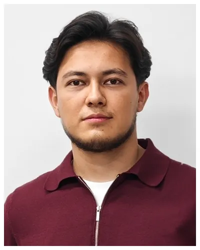 Portrait of a young man with dark hair wearing a maroon zip-up shirt against a plain background.