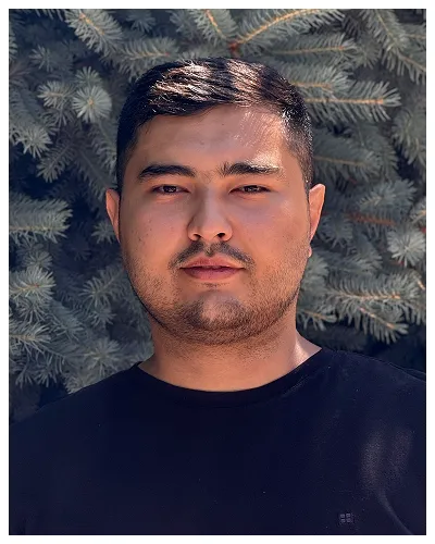 Portrait of a young man with short dark hair wearing a black shirt, standing outdoors against green pine tree branches.