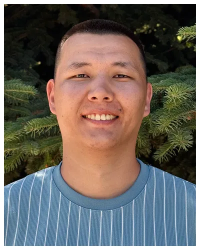 Portrait of a smiling man wearing a blue striped shirt with green pine trees in the background.