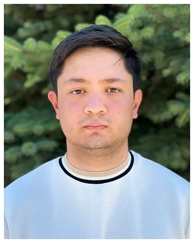 Portrait of a young man with short dark hair wearing a white shirt, against a green pine tree background.