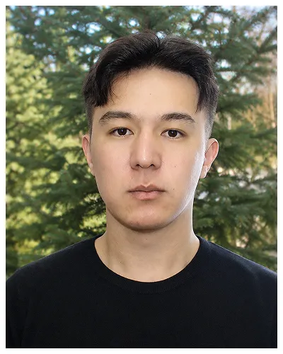 Portrait of a young man with short dark hair wearing a black shirt, standing outdoors with green trees in the background.