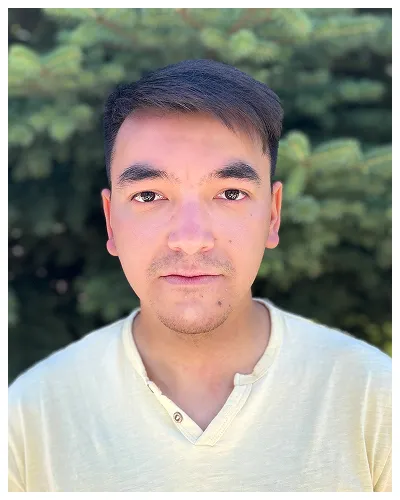 Portrait of a young man with short dark hair wearing a light yellow shirt, outdoors with blurred green foliage background.