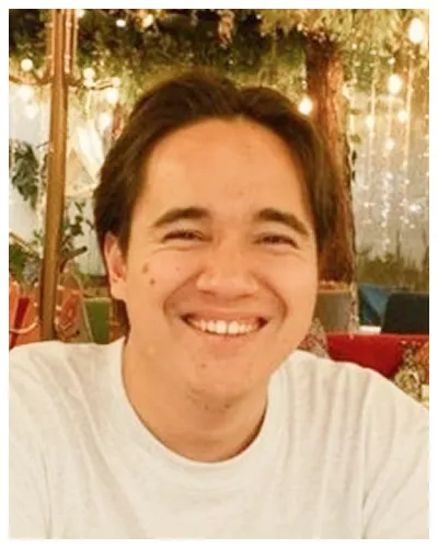 Portrait of a smiling young man in a white shirt with warm ambient lighting and greenery in the background.