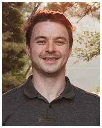 Portrait of a smiling young man with short brown hair wearing a dark polo shirt outdoors.