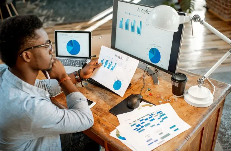 Man Sitting at Desk Looking at Analytics on Computer and Documents