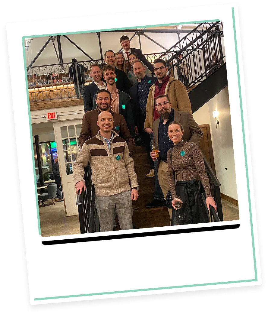 Group of eleven adults posing on indoor wooden staircase, dressed in business casual attire, some holding drinks.