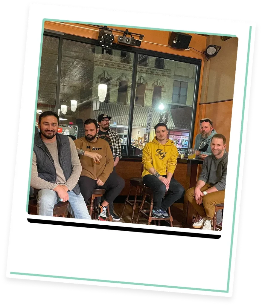 Six casually dressed men sitting on bar stools inside a pub with large windows showing a nighttime street view.