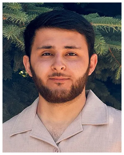 Portrait of a young man with a beard, wearing a beige shirt, standing outdoors by green pine branches.