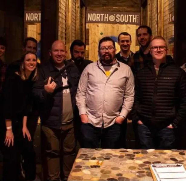 Group of eight adults standing indoors in front of wooden axe-throwing targets labeled "NORTH SOUTH" under warm lighting.