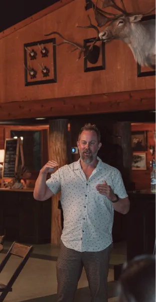 Man speaking indoors with mounted deer head and rustic wooden decor in background.