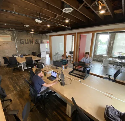 Open-plan office with exposed wooden beams, multiple desks, three people working on laptops.