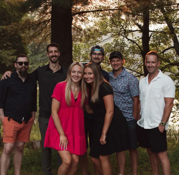 Group of seven adults posing outdoors near trees and water, dressed in casual summer clothing.