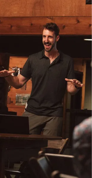 Man in black polo shirt speaking animatedly in a warmly lit wooden interior.