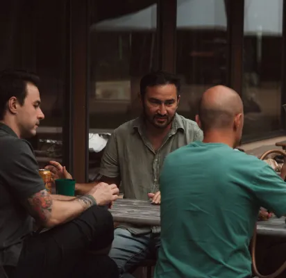 Three men engaged in conversation around a wooden table in an indoor setting.
