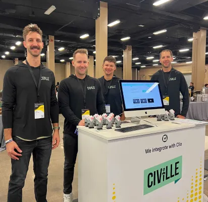 Four men stand behind a white exhibition booth with a computer monitor and promotional items, wearing black Civille-branded shirts.