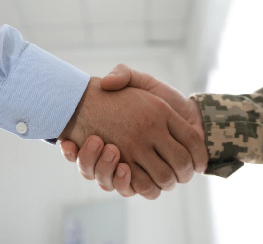 Close-up handshake between a civilian in a suit and a person in military camouflage uniform.