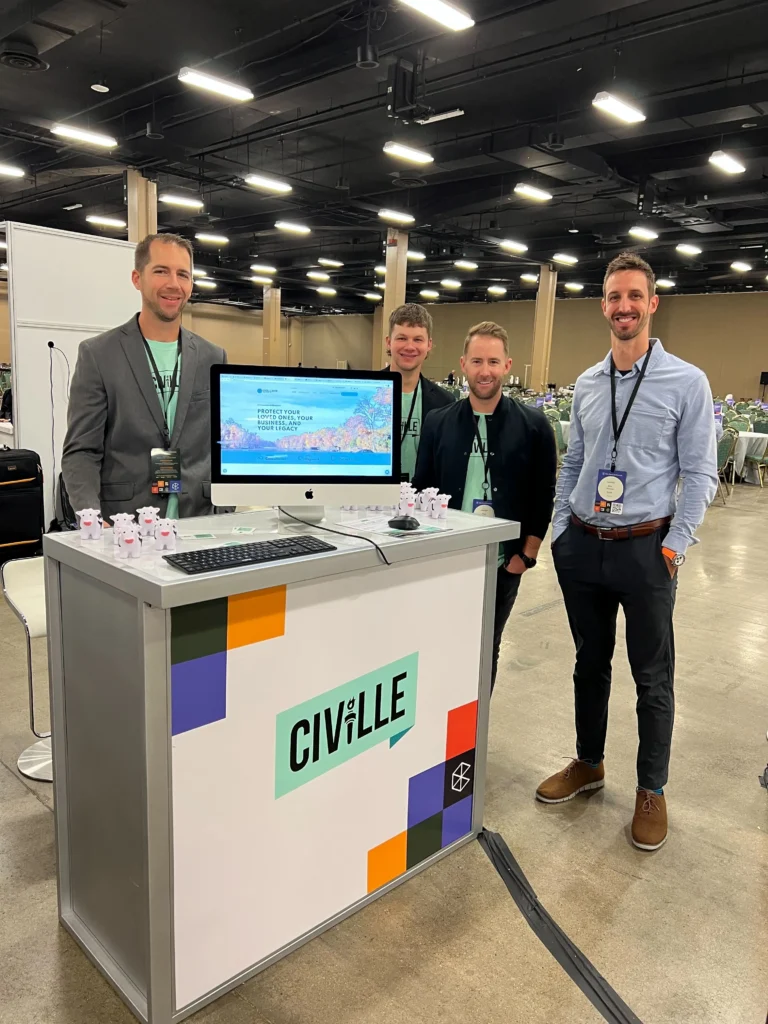 Four men standing behind a Civille branded booth with a computer monitor displaying a website in a large conference hall.