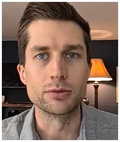 Close-up of a young man with blue eyes and short brown hair, indoors with a lit table lamp in the background.