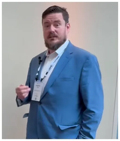 Man with beard wearing blue suit and white shirt, speaking with conference badge visible.