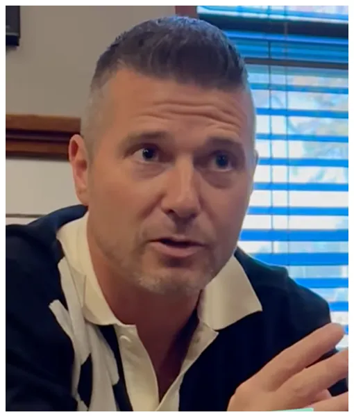 Middle-aged man with short hair speaking indoors, wearing a black and white collared shirt, blue window blinds in background.