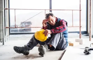 Construction worker sitting on the floor holding a yellow hard hat, appearing distressed.