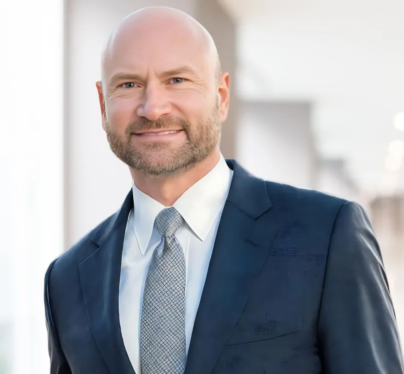 Professional middle-aged bald man with beard wearing navy suit and patterned tie, smiling.
