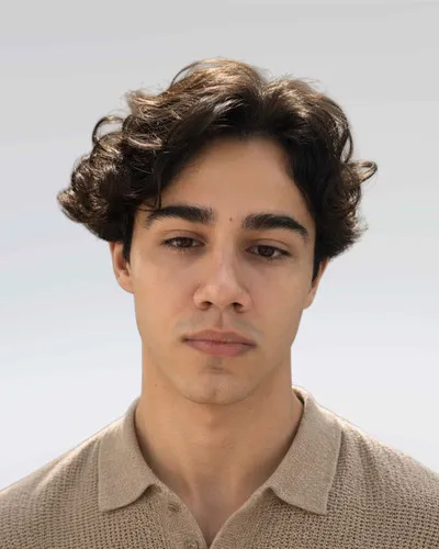 Portrait of a young man with curly dark hair wearing a beige collared shirt.